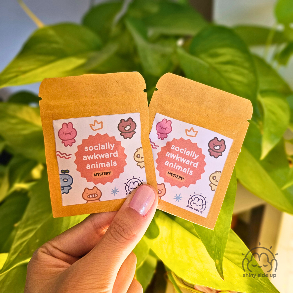 Two small orange packages with a label reading 'socially awkward animals' held against a green leafy background.